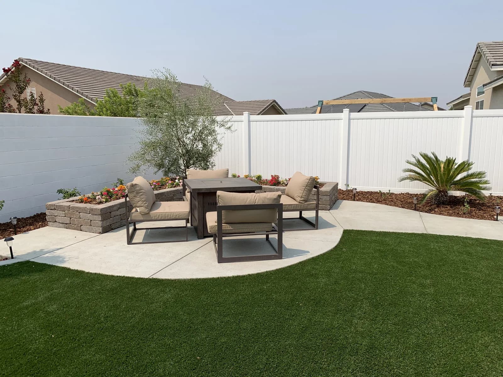 A fire pit with four chairs on a round paver patio, set in artificial grass beside a white fence