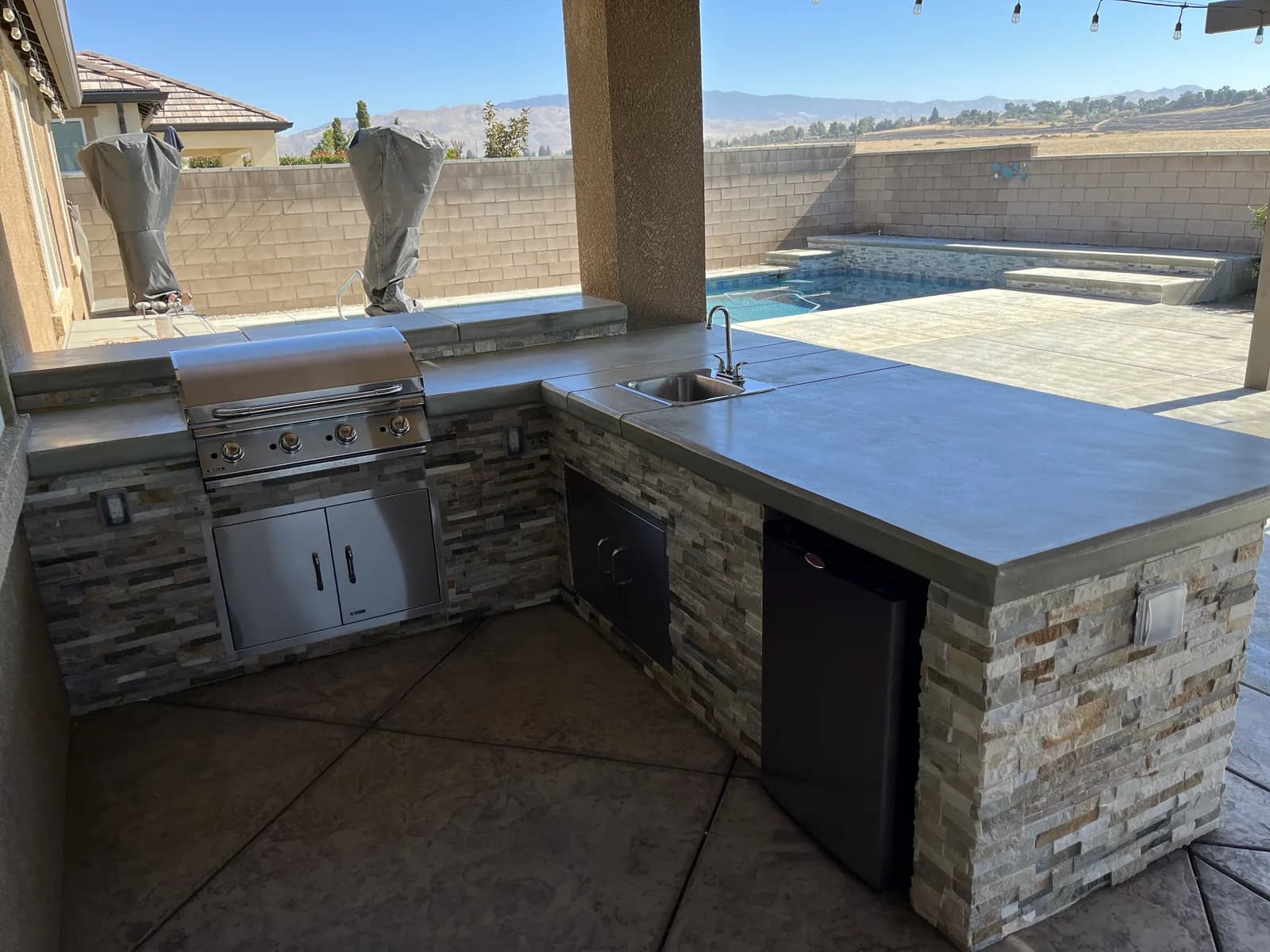 An outdoor kitchen with a stone base and concrete counter under a patio cover, a pool behind it