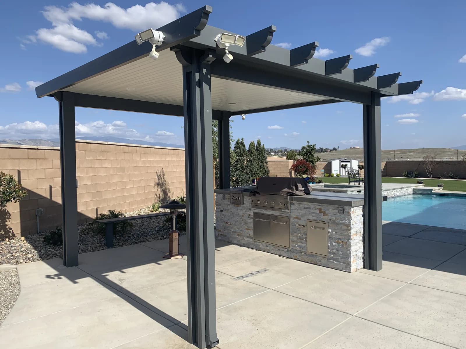 A pergola with an outdoor kitchen beside a pool, open land beyond the block wall