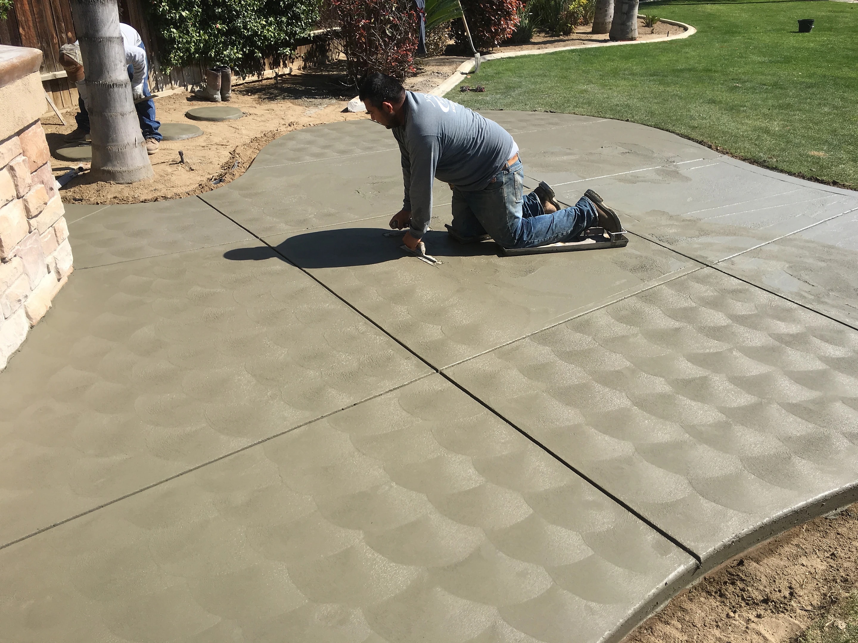 A crew member on his knees finishing a freshly poured concrete patio by hand, beside a lawn