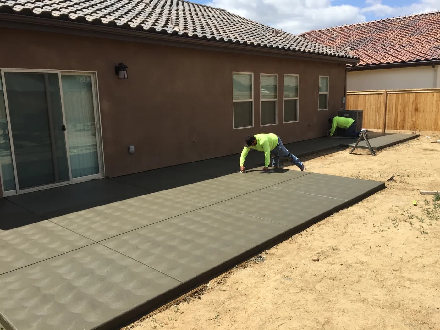 Two crew members finishing a stamped concrete patio behind a house, the yard still bare dirt