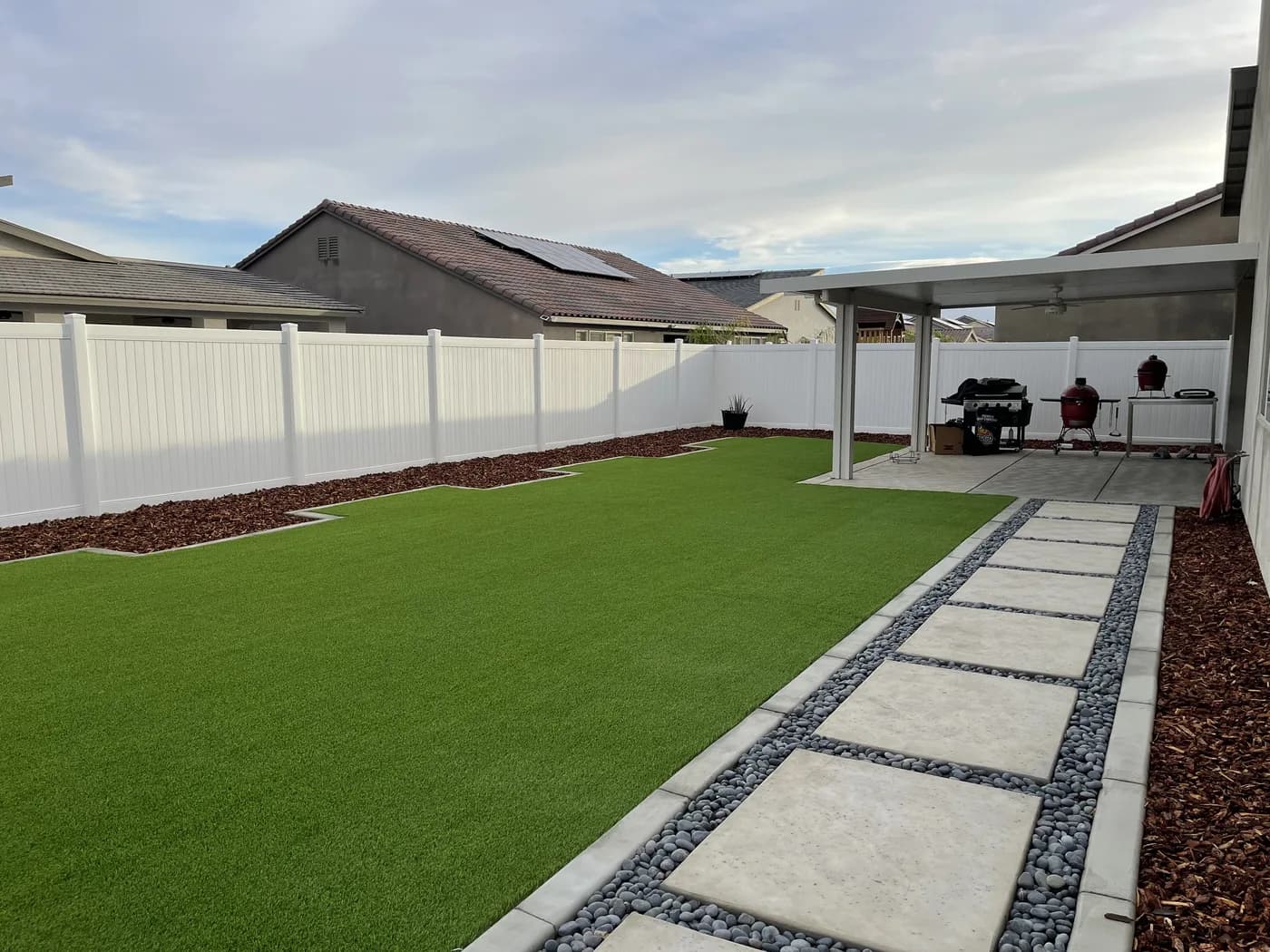 Concrete stepping pads set in pebbles along a turf side yard, with a patio cover behind