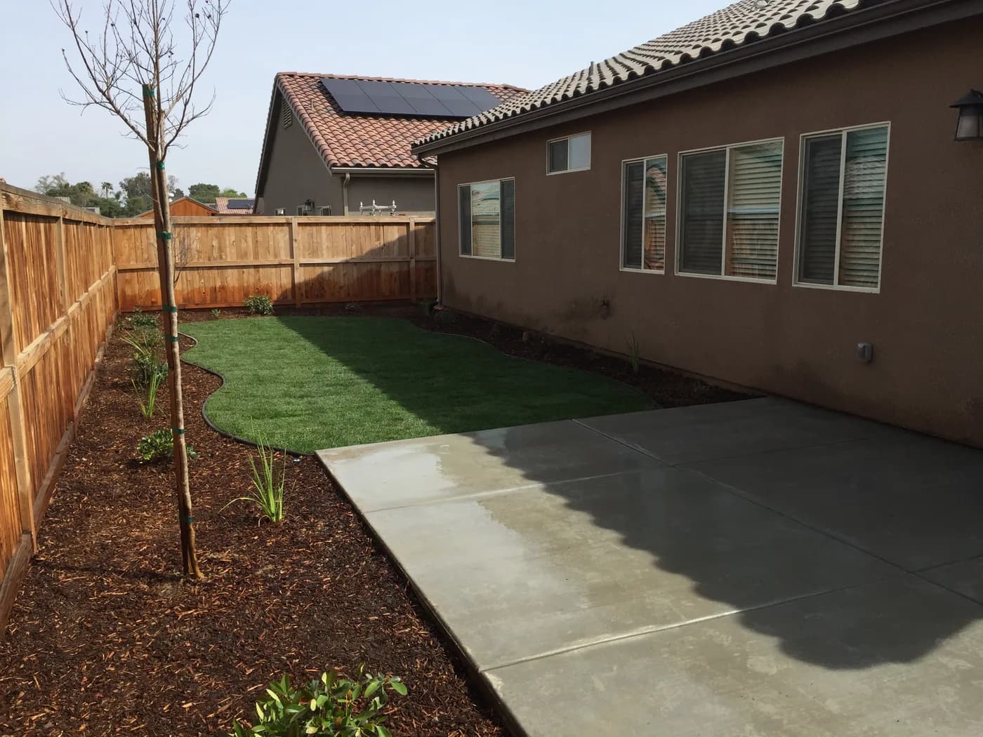 Broom-finished concrete patio beside a small turf lawn, a young tree and a bark bed