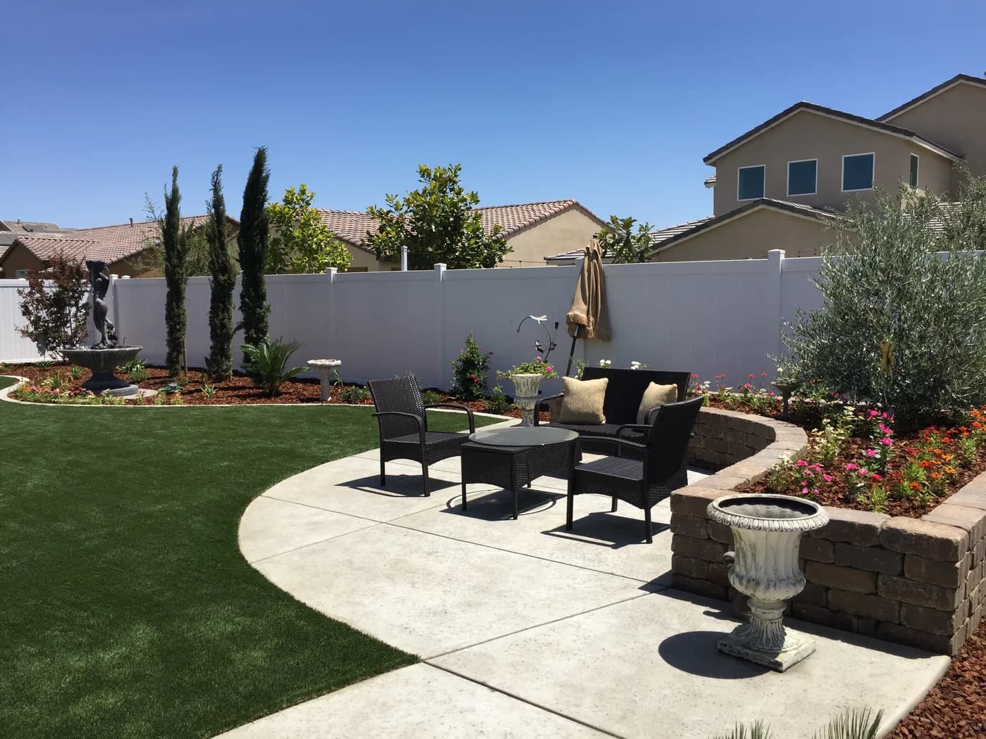 Curved concrete patio with wicker seating, a block planter full of flowers, turf and cypress trees
