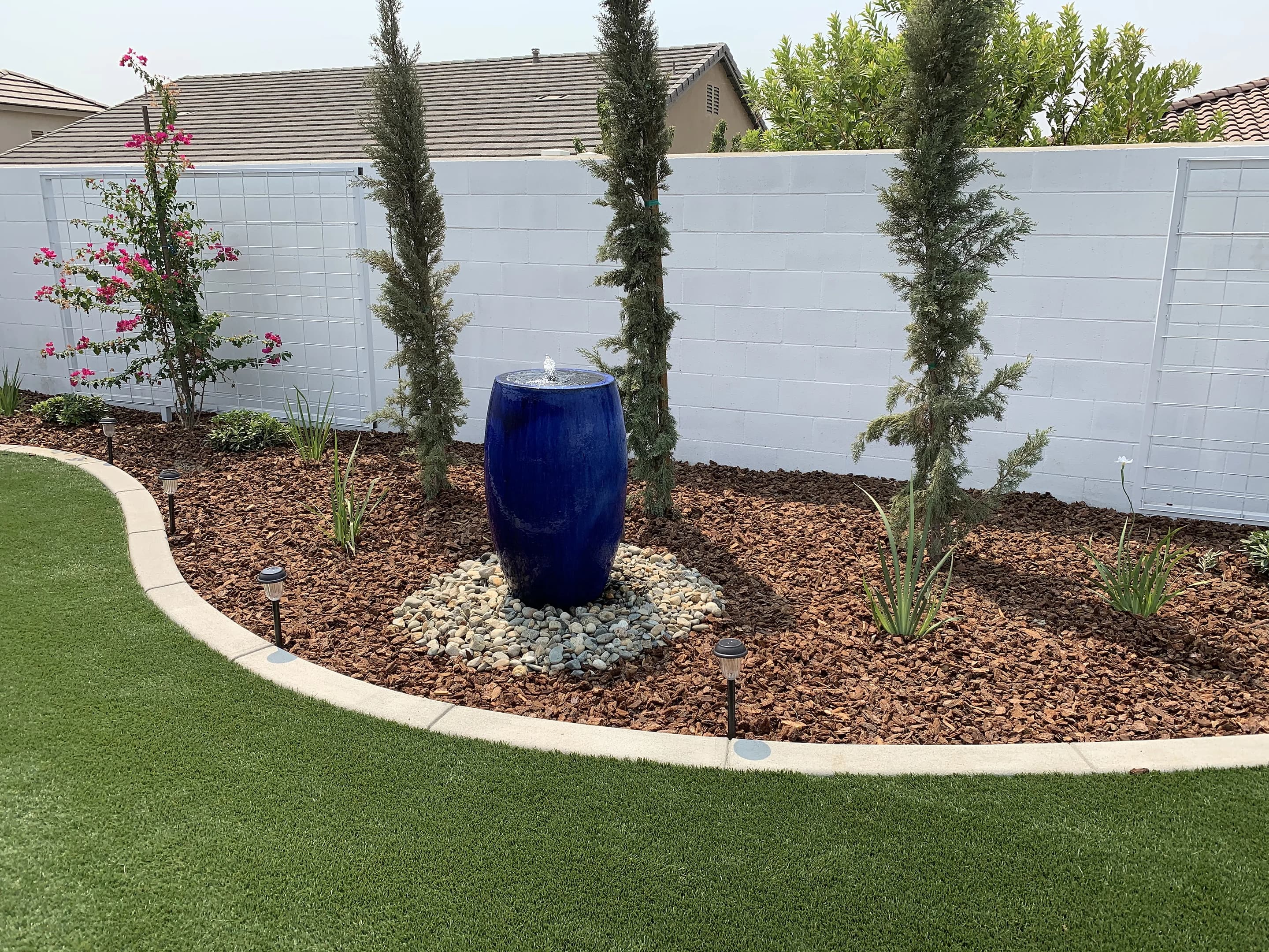 Blue ceramic fountain among Italian cypress, river rock and bark beside a lawn with path lights