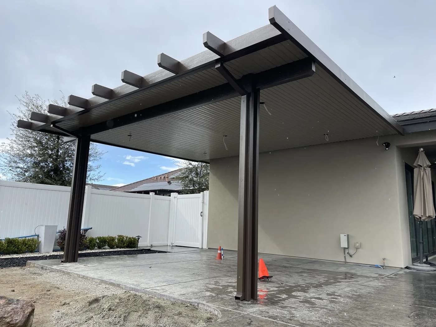 Dark patio cover standing on a freshly finished concrete slab, with cones still out
